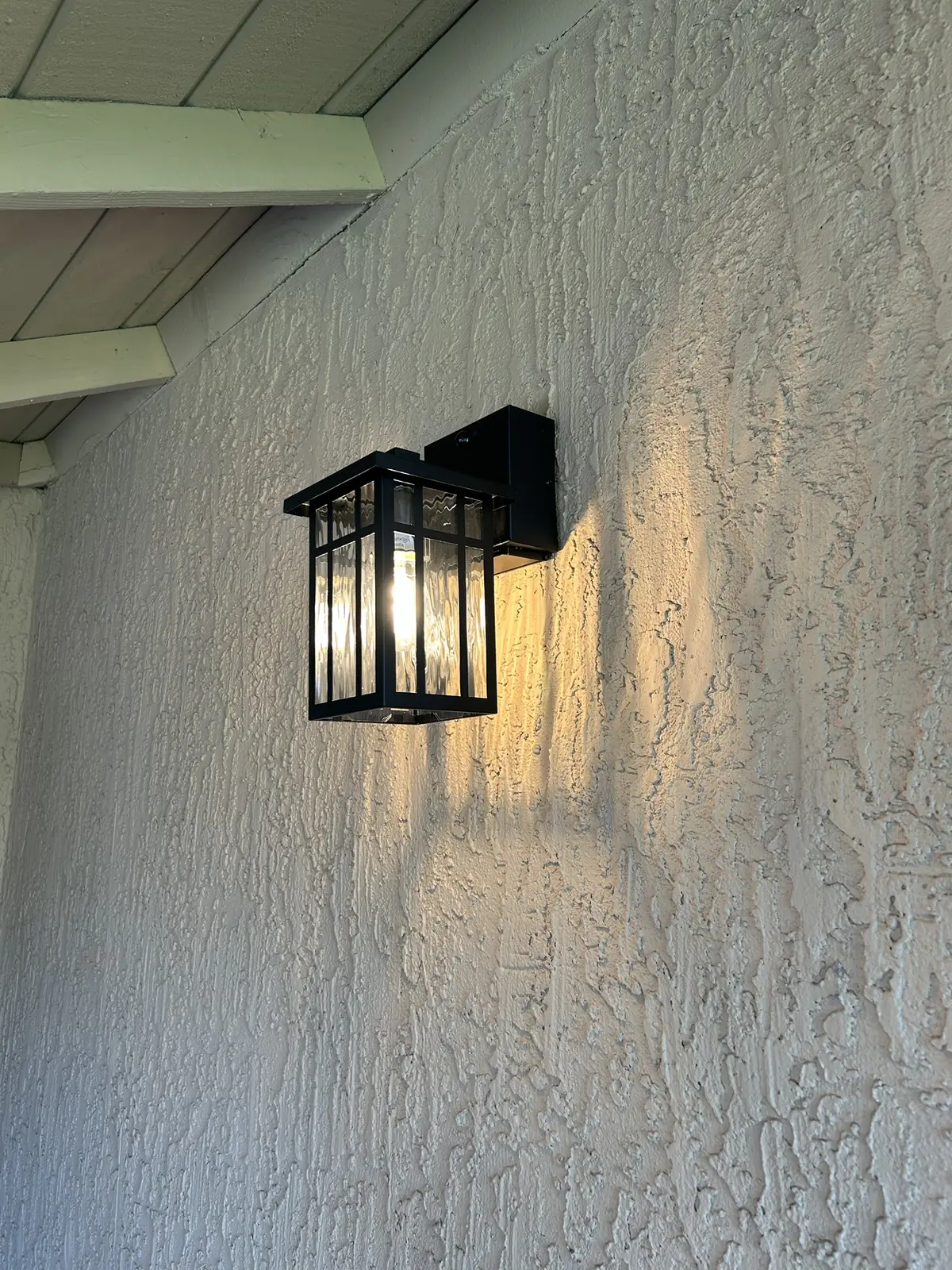 Black craftsman-style wall lantern glowing warm against a textured stucco wall