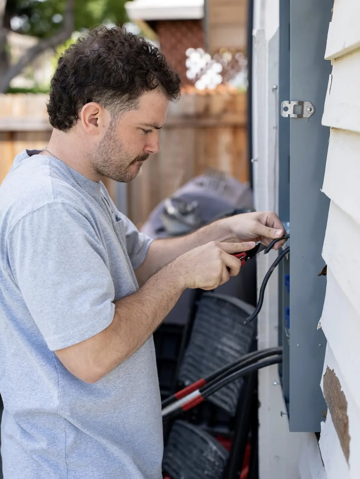 Handyman in a light blue t-shirt stripping wire at an outdoor electrical panel